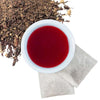 Brewed hibiscus ginger herbal tea with two tea bags and loose contents of tea bags in upper left hand corner